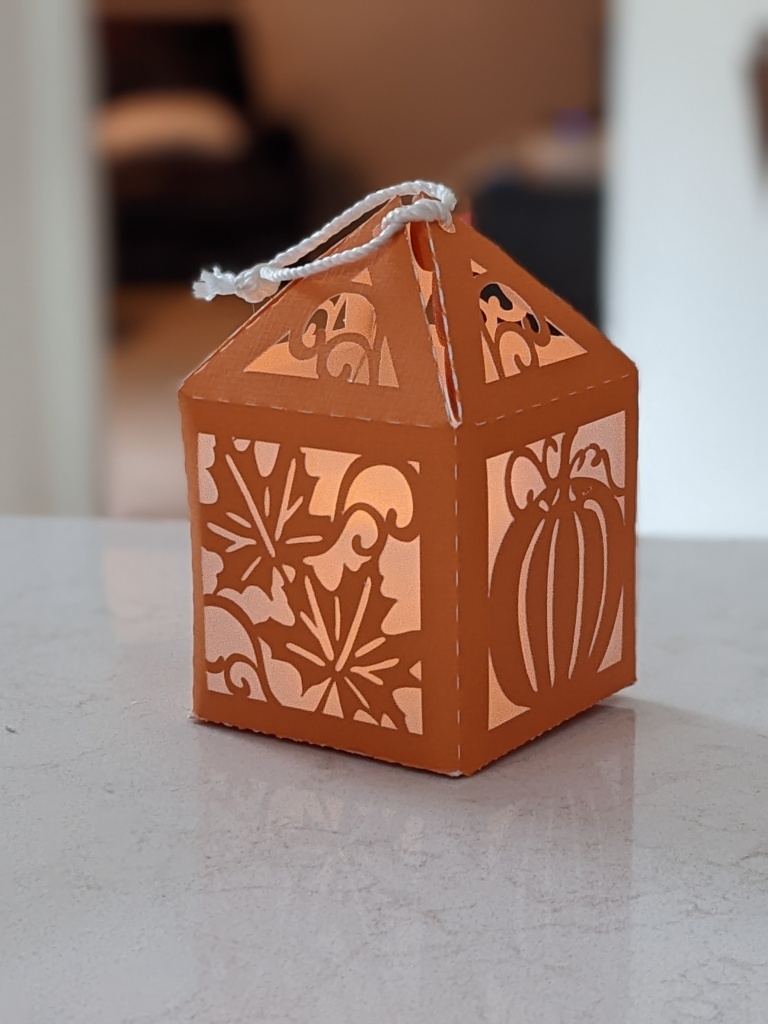 A photo of a cricut-cut paper lantern. The paper is brown and depicts maple leaves and a pumpkin, with the negative space covered by transparent vellum. A light is visible from within.