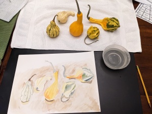 Five gourds have been laid out on a white towel. Below it is a watercolor of those gourds. There is a yellow gourd with green stripes sitting up, a white-yellow gourd on its side facing right. An orange gourd facing up. A small ball-like yellow and green striped gourd facing left. A larger gourd with a green and yellow striped bottom and orange top, laying on its side facing left.
